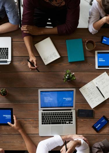Zu sehen ist ein Tisch mit Arbeitsutensilien wie Laptops, Stiften und Ipads, um den Menschen, die nicht erkennbar sind, herumsitzen
